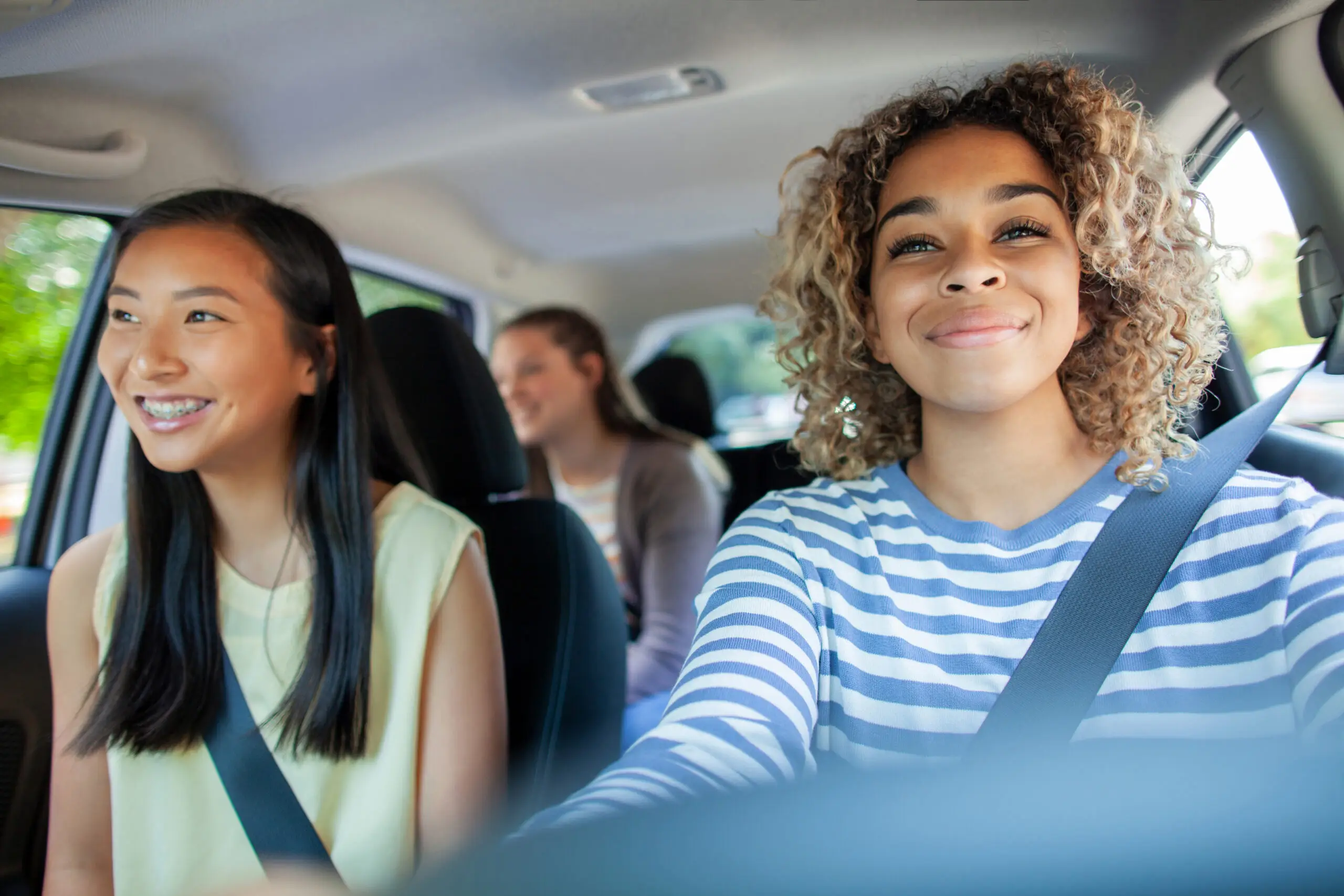 Three young women smiling and wearing seatbelts inside a car, one in the driver's seat.
