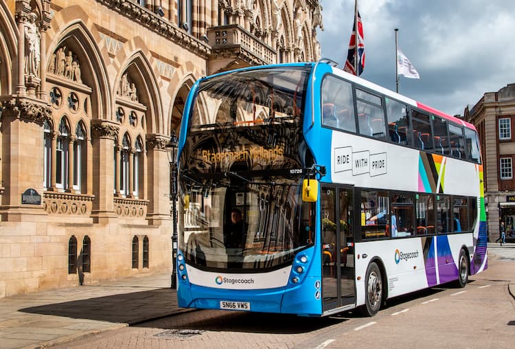 A blue and white double-decker Stagecoach bus with a "Ride with Pride" message and colorful geometric patterns drives down a street in front of a large, ornate stone building.