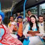 A group of people in elaborate costumes, including a clown, a prince, and a princess, are seated on a bus, with golden glitter effects scattered around them. GO Travel Solutions to host Better Transport Week webinars