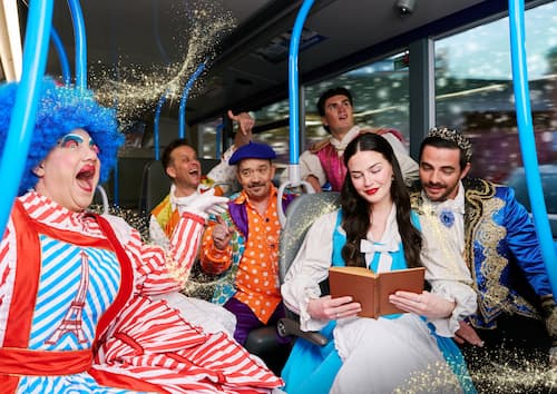 A group of people in elaborate costumes, including a clown, a prince, and a princess, are seated on a bus, with golden glitter effects scattered around them. GO Travel Solutions to host Better Transport Week webinars