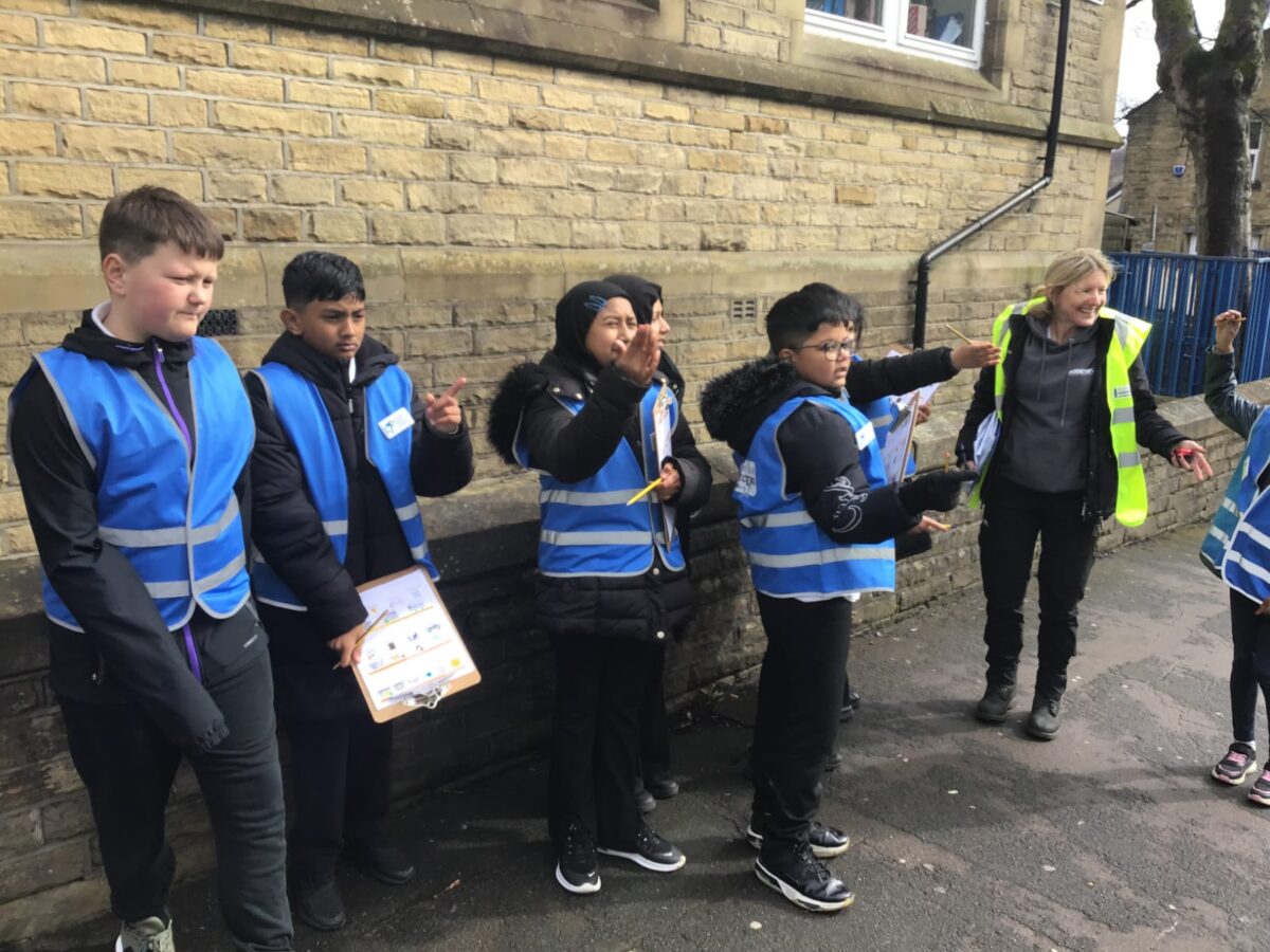 Group of children wearing blue safety vests standing outside near a brick wall, engaged in an outdoor activity with an adult in a yellow vest. Holycroft Primary School, Bradford