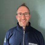 A smiling man wearing glasses and a navy blue quarter-zip sweatshirt with the "MODESHIFT SUPPORTING EVERYDAY JOURNEYS" logo on the front.
