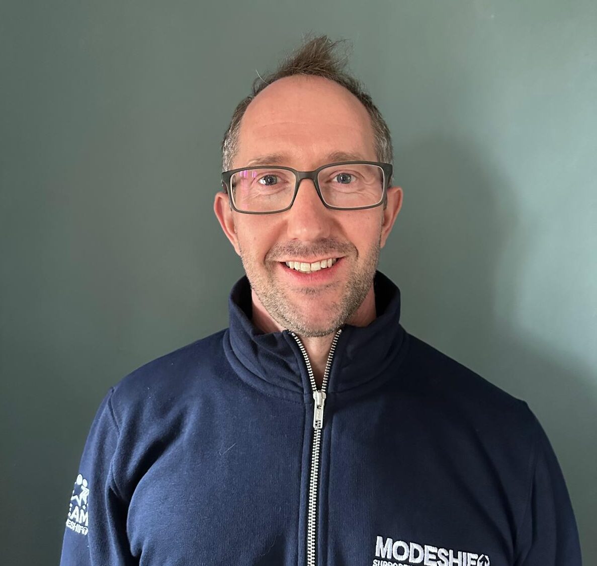 A smiling man wearing glasses and a navy blue quarter-zip sweatshirt with the "MODESHIFT SUPPORTING EVERYDAY JOURNEYS" logo on the front.