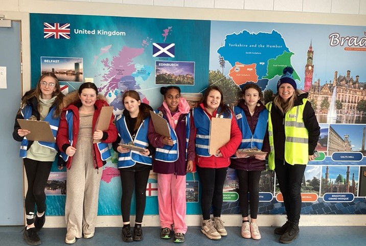 A group of seven young people, most wearing blue safety vests, stand in front of a colorful mural depicting maps of the United Kingdom and Yorkshire. Several of the young people hold clipboards.