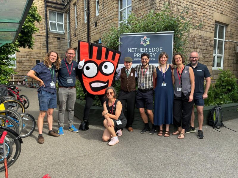 Group of adults and a person in a large red hand mascot costume posing outside Nether Edge Primary School on a sunny day.