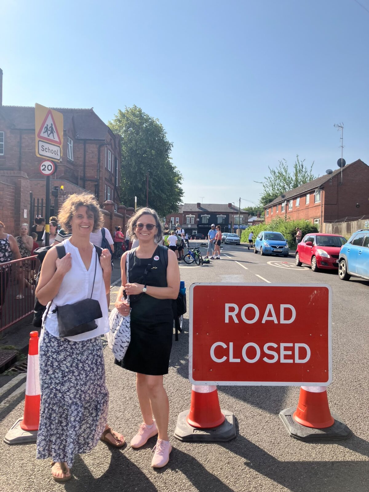 Two women stand on a street next to a red "ROAD CLOSED" sign, with a school building visible in the background and several parked cars further down the road.