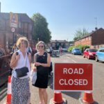 Two women stand on a street next to a red "ROAD CLOSED" sign, with a school building visible in the background and several parked cars further down the road.