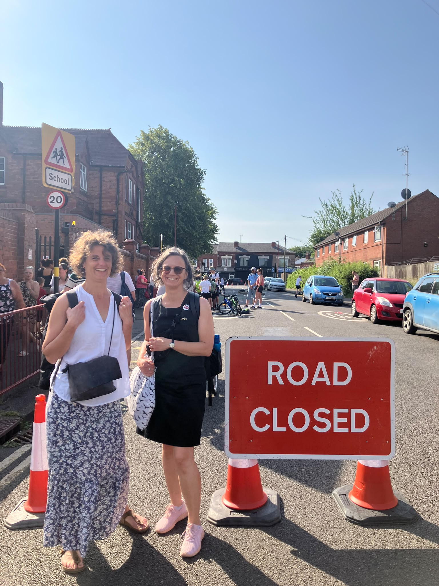 Two women stand on a street next to a red "ROAD CLOSED" sign, with a school building visible in the background and several parked cars further down the road.