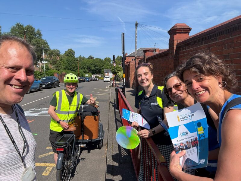 Group of people outdoors near a brick wall, one person in a yellow safety vest and helmet with a cargo bike, others holding informational pamphlets and a colorful disc under a clear blue sky.