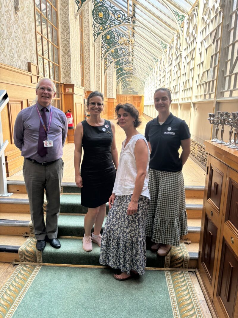 Four adults standing on carpeted stairs inside a sunlit hallway with large windows and ornate ceiling decorations.