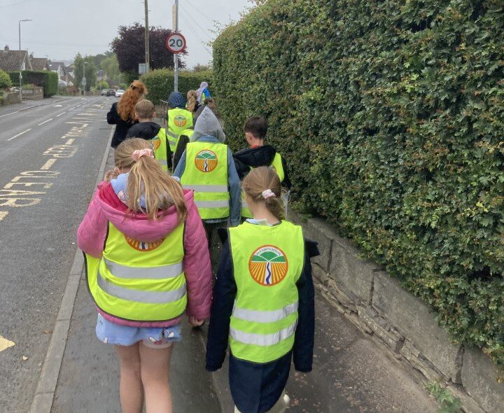Old Earth Primary School Children wearing bright yellow high-visibility vests with a green and orange circular logo walk single file along a pavement next to a dense green hedge.
