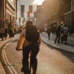 A person wearing a backpack rides a bicycle down a city street during golden hour, with buildings lining the road and a crowd of pedestrians in the background. Survey is being conducted by the Transport Planning Society (TPS)