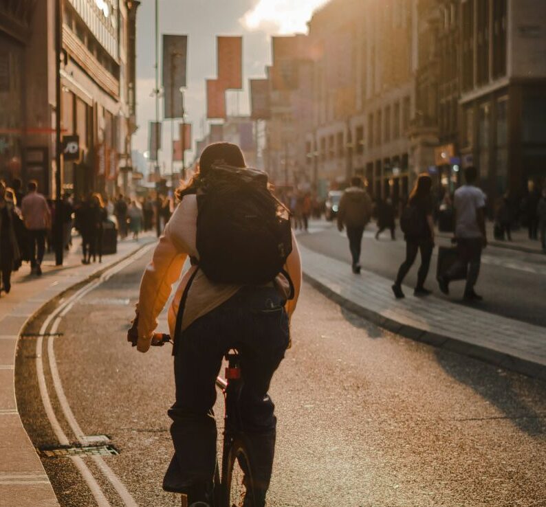A person wearing a backpack rides a bicycle down a city street during golden hour, with buildings lining the road and a crowd of pedestrians in the background. Survey is being conducted by the Transport Planning Society (TPS)