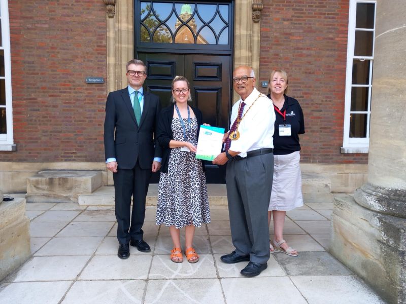 Four people stand on a stone patio in front of a red brick building. A man in a dark suit and green tie stands on the left, looking towards the camera. Next to him, a woman in a patterned dress and black cardigan holds a certificate. To their right, a man wearing a ceremonial chain of office over a white shirt and grey trousers holds the certificate. Behind him, a woman in a black polo shirt and light-colored skirt looks towards the camera.