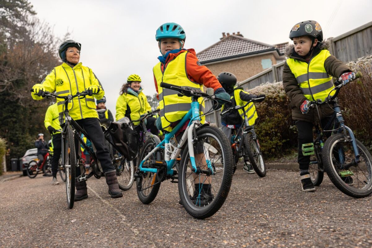 Group of children wearing helmets and high-visibility vests riding bicycles on a suburban street.