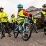 Group of children wearing helmets and high-visibility vests riding bicycles on a suburban street.
