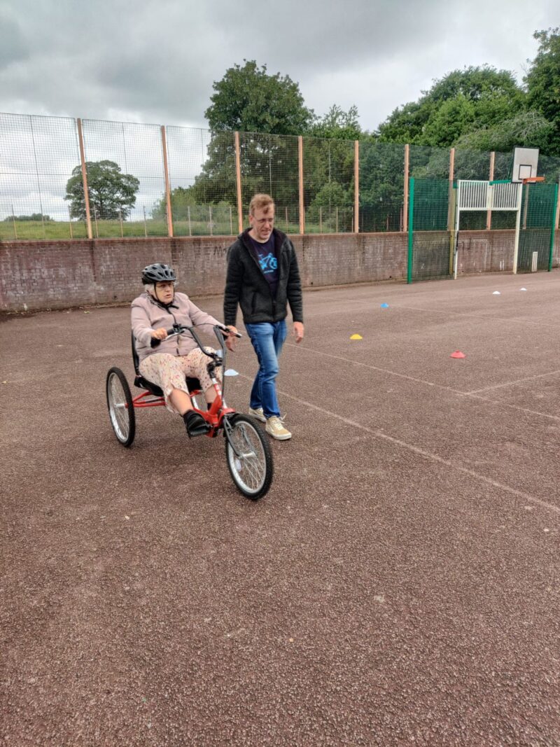 A person wearing a helmet rides a red adaptive tricycle on an asphalt court while another person stands beside them.