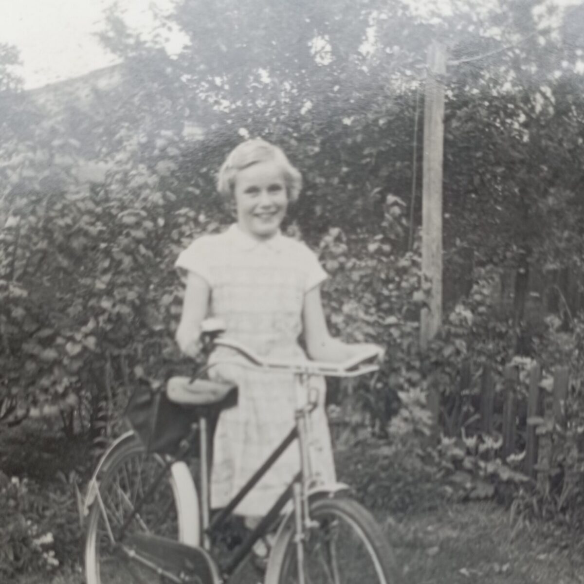 A young girl smiles as she stands next to a bicycle in a grassy yard, with dense foliage and a wooden fence in the background.