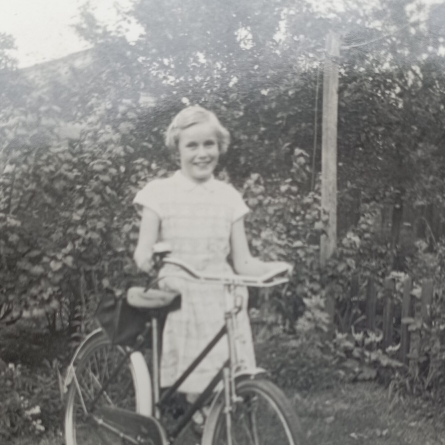 A young girl smiles as she stands next to a bicycle in a grassy yard, with dense foliage and a wooden fence in the background.