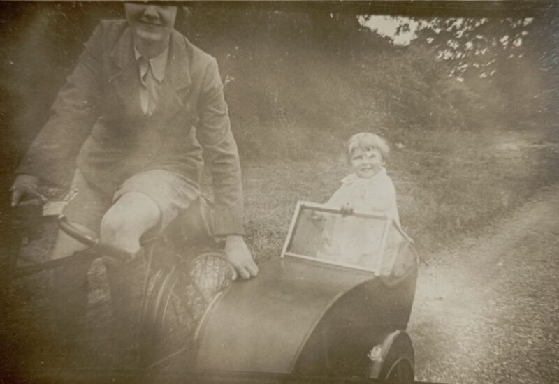 A black and white photograph - child in a side car with an adult on a bicycle.