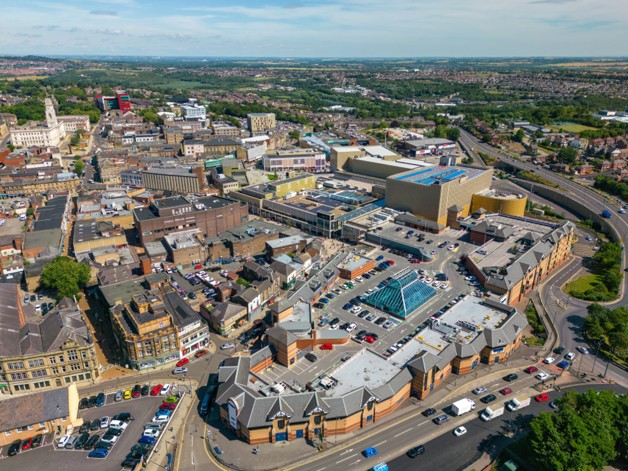 BetterPoints launches physical activity rewards programme across Barnsley. Aerial view of a bustling town center with a large shopping complex surrounded by streets and parked cars under a partly cloudy sky.