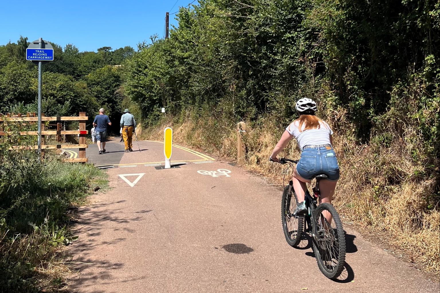 A cyclist wearing a helmet and denim shorts rides on a paved path bordered by trees and vegetation, with two walkers ahead and a sign indicating "TRAIL REJOINS CARRIAGEWAY."