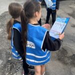 Four children wearing blue high-visibility vests examine a checklist on a clipboard outdoors, with cars parked in the background.