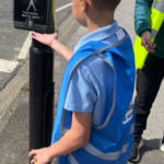 A child wearing a blue safety vest and a school uniform reaches out towards a pedestrian crossing signal showing a red standing figure, indicating "Don't Walk."