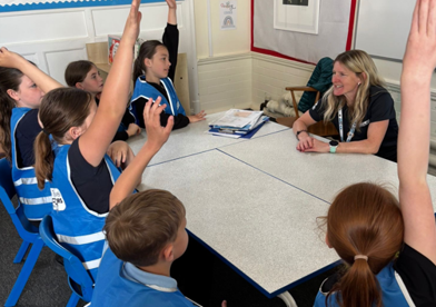 Children wearing blue "Active Travel Inspectors" vests sit around a table with their hands raised, facing a smiling adult.