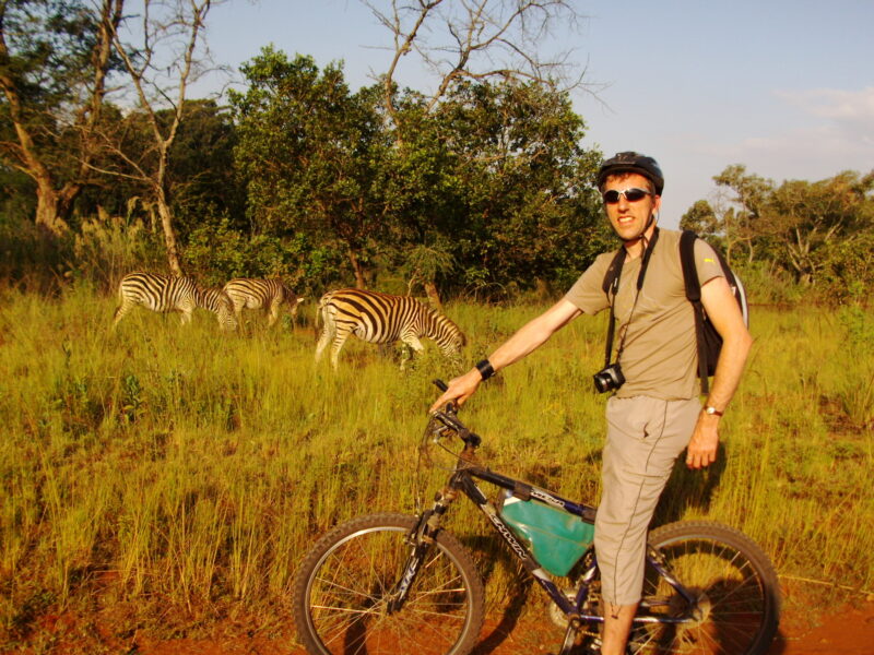 A cyclist in a helmet and sunglasses pauses on a dirt path with a bicycle, looking towards the camera, as several zebras graze in the tall, dry grass of a savanna landscape under a clear sky.