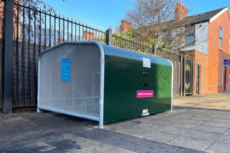 A dark green, enclosed, and covered bike storage unit with a perforated metal front, situated on a paved sidewalk next to a black metal fence.