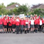 A diverse group of children in red school uniforms and a few adults in casual attire and high-visibility vests pose for a photo outdoors, holding up signs, with one sign featuring a bicycle illustration.