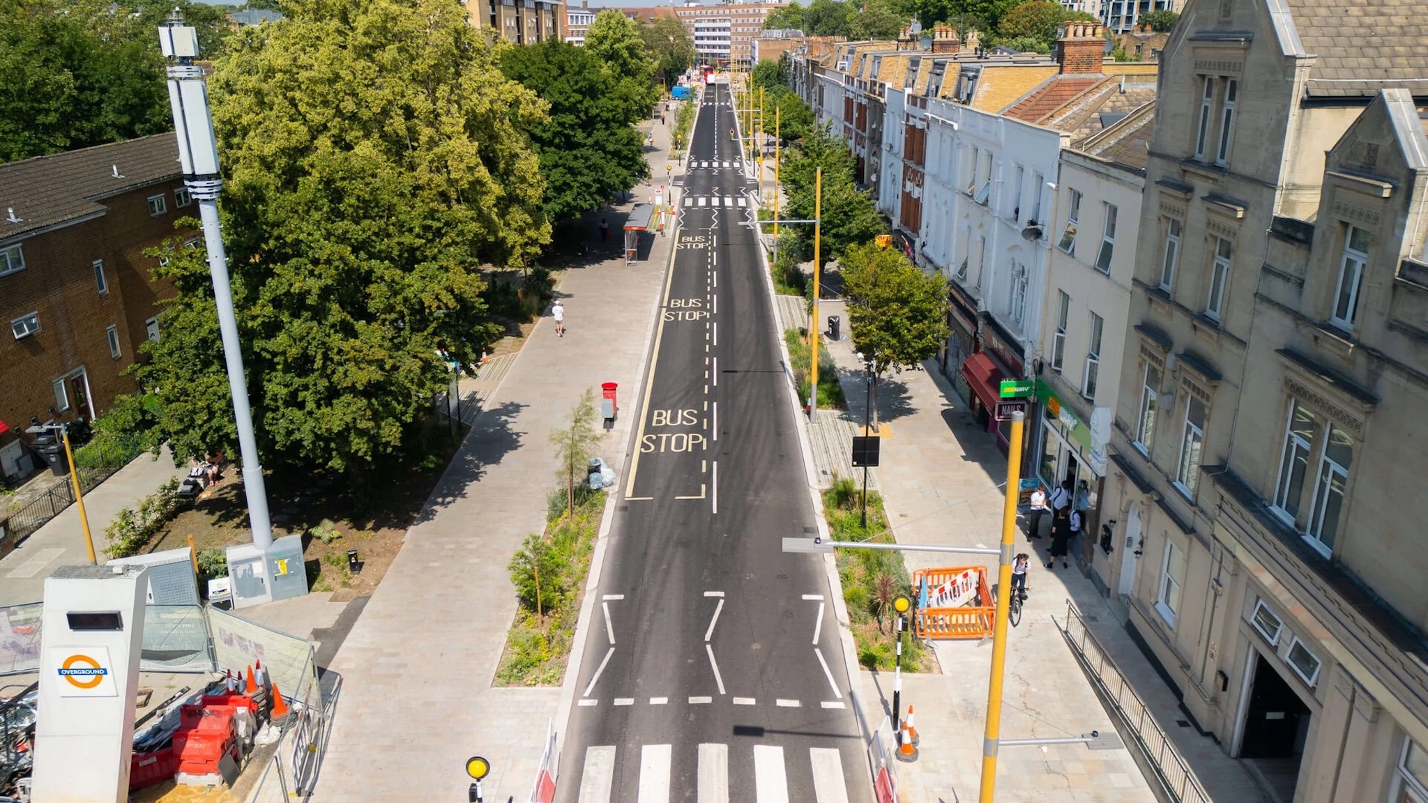 Aerial view of a newly paved street with pedestrian crossings, bus stop markings, and trees lining the sidewalks, bordered by buildings on one side and residential buildings and greenery on the other.