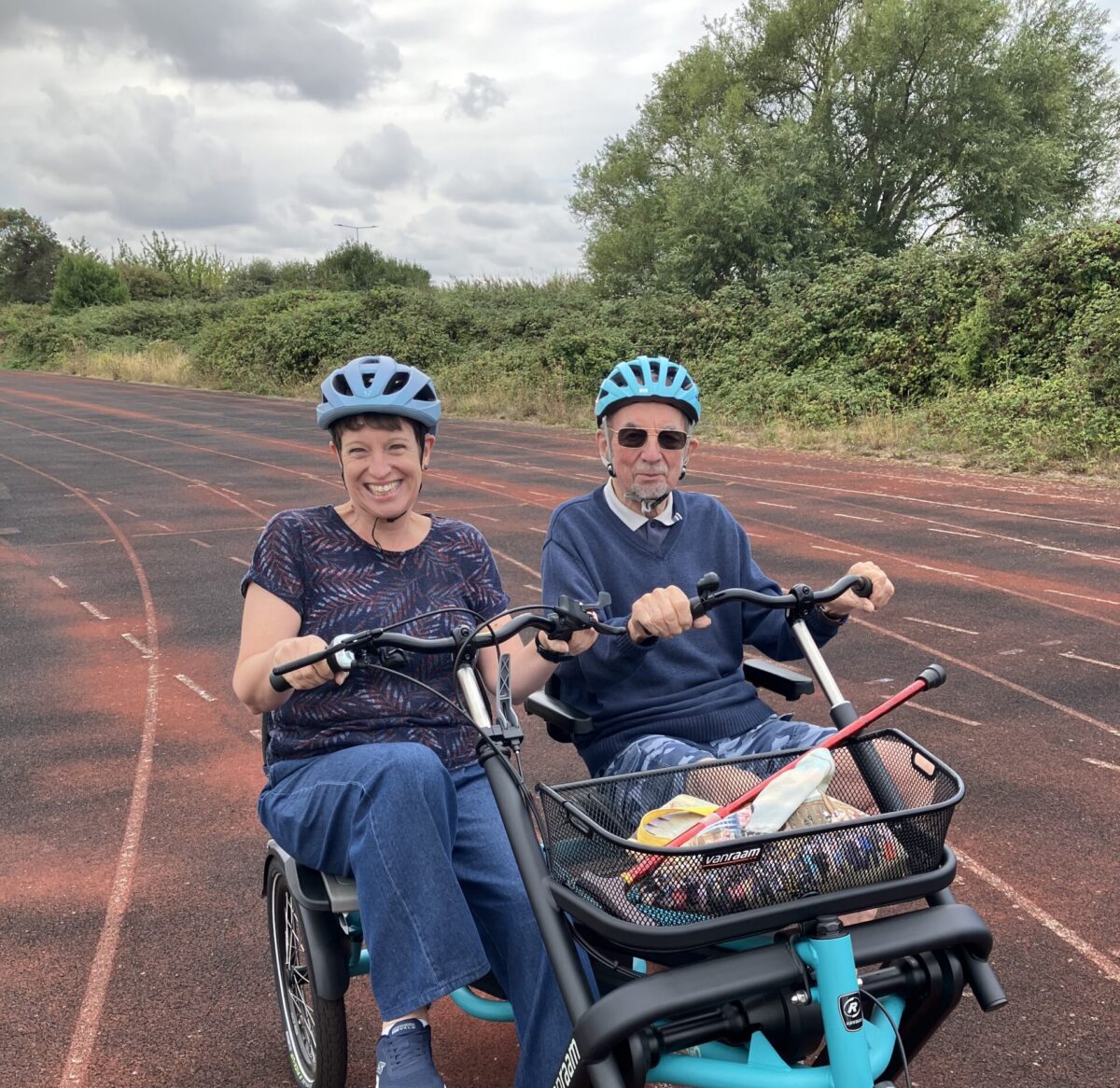 Two people wearing blue helmets ride matching teal recumbent tricycles on a reddish-brown track. Mode Shift Month activity ideas.