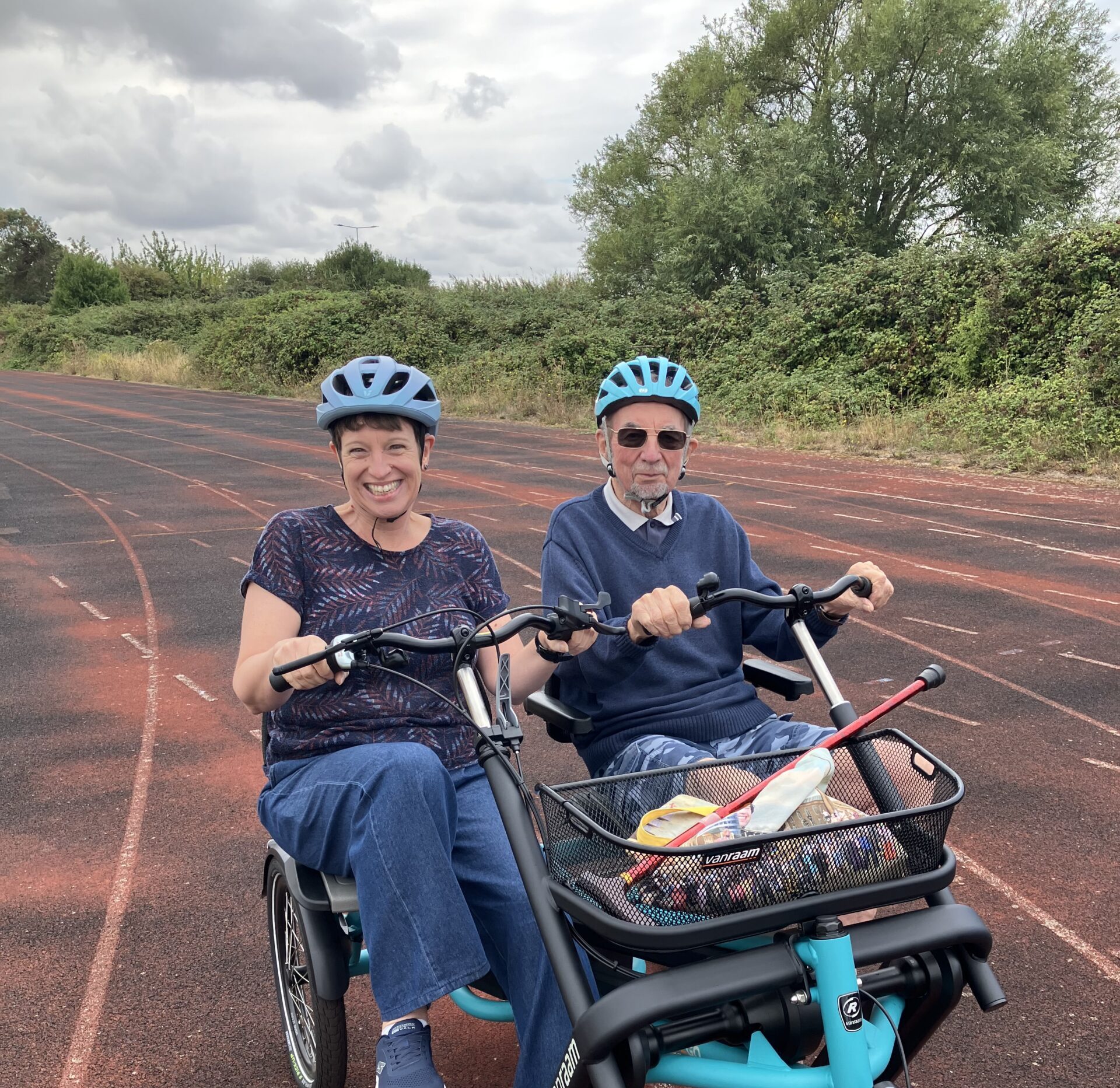 Two people wearing blue helmets ride matching teal recumbent tricycles on a reddish-brown track. Mode Shift Month activity ideas.