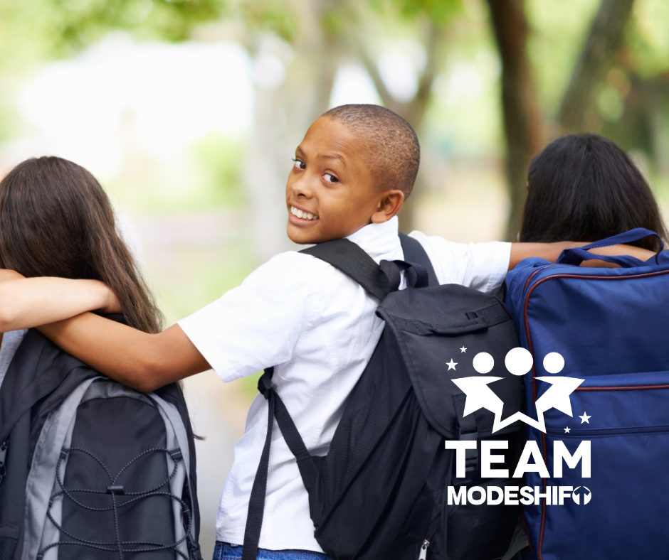 Three children walk together outdoors with backpacks, one child in the center looks back at the camera and smiles. A white logo with text "TEAM MODESHIFT" is overlaid on the image.