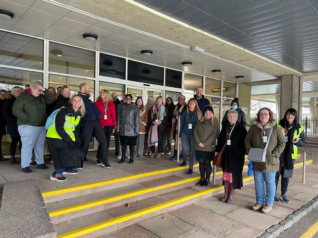 A diverse group of people stands on steps outside a modern building with a covered entrance.