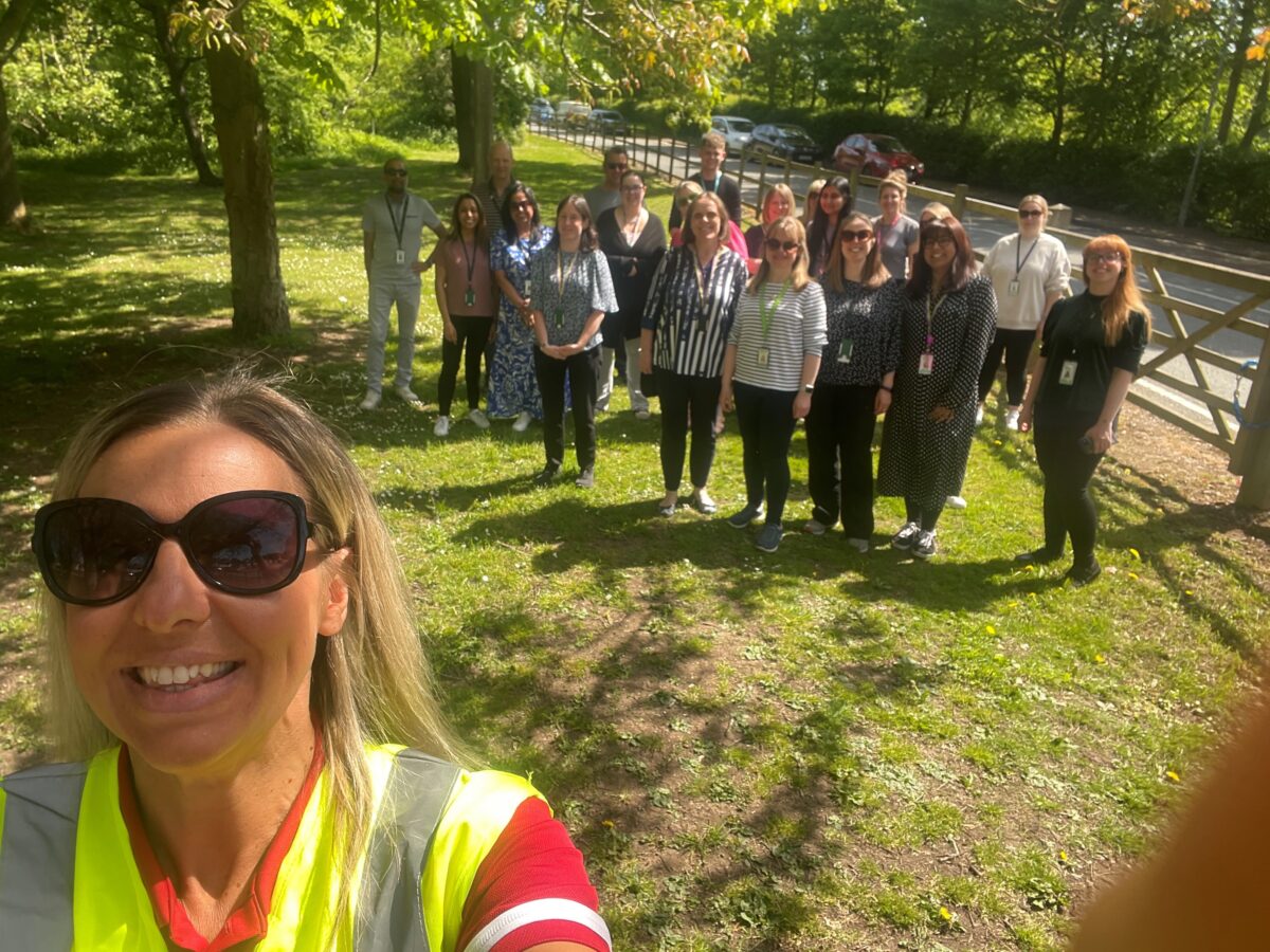 A group of people in casual clothing stand on a grassy area with trees and a fence in the background, with a woman in the foreground wearing sunglasses and a high-visibility vest. Case Study: Choose How You Move Wellbeing Walks, Leicestershire County Council