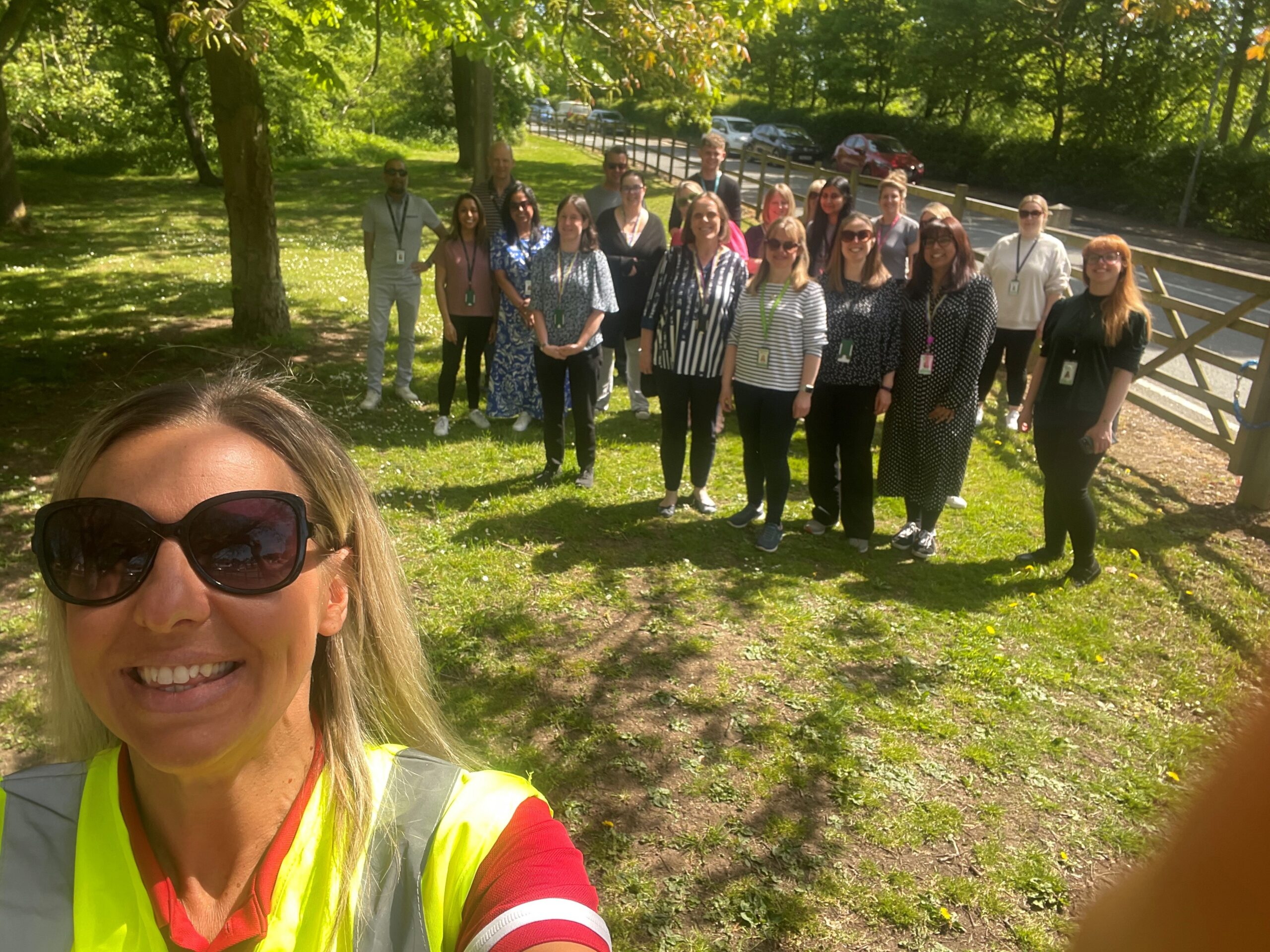 A group of people in casual clothing stand on a grassy area with trees and a fence in the background, with a woman in the foreground wearing sunglasses and a high-visibility vest. Case Study: Choose How You Move Wellbeing Walks, Leicestershire County Council
