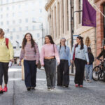 A diverse group of people walk down a city street past buildings and a pedestrian zone sign.