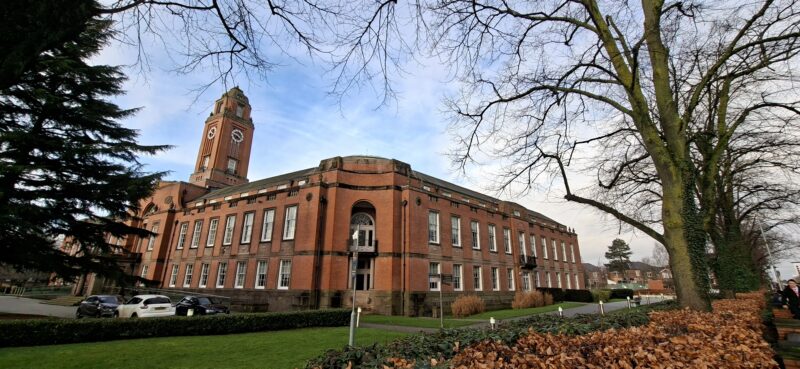 A grand brick building with a tall clock tower, seen from a low angle with bare tree branches framing the sky.