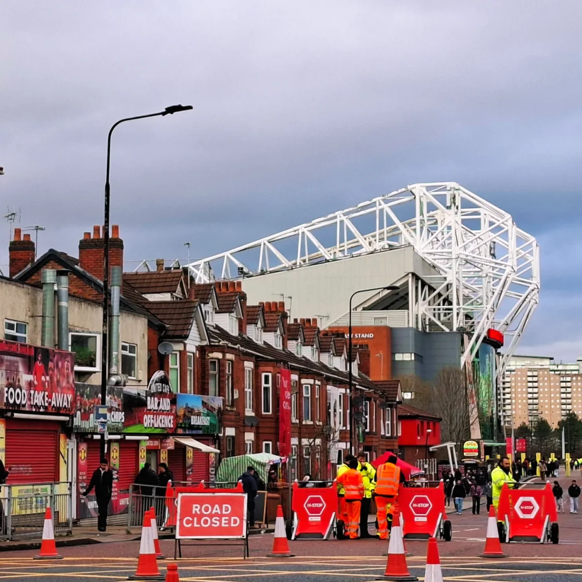 Street scene in front of a large stadium with a white lattice roof, featuring shops and several workers in high-visibility vests setting up road closure barriers.