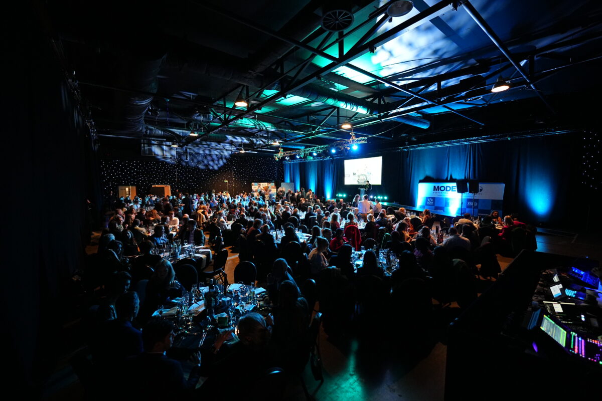 A large, dimly lit event hall filled with round tables of guests seated for a formal dinner, with a stage at the front featuring a large screen displaying a presentation and a "MODESHIFT" logo.
