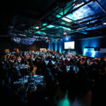 A large, dimly lit event hall filled with round tables of guests seated for a formal dinner, with a stage at the front featuring a large screen displaying a presentation and a "MODESHIFT" logo.