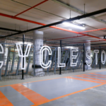 Large white letters spell out "CYCLE STORE" in front of a fenced-off area with bicycle racks in a parking garage. Turvec photo credit.