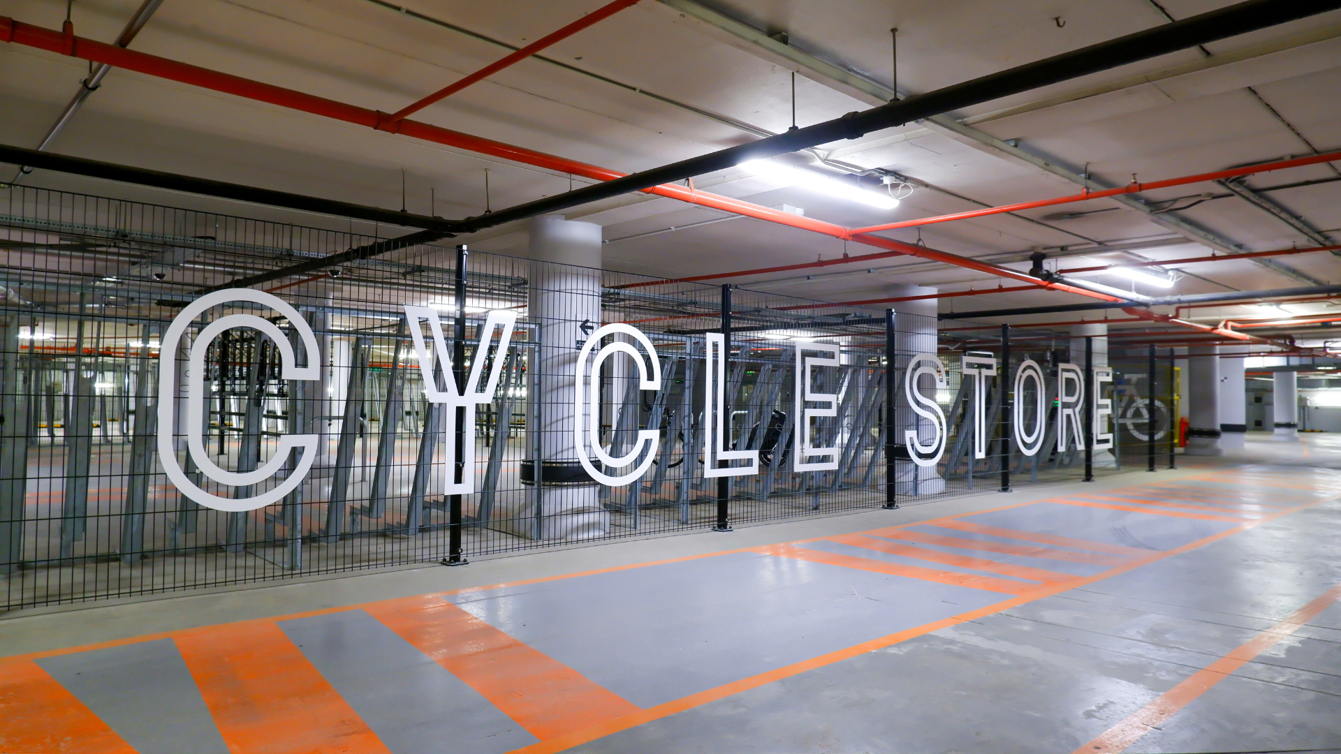 Large white letters spell out "CYCLE STORE" in front of a fenced-off area with bicycle racks in a parking garage. Turvec photo credit.