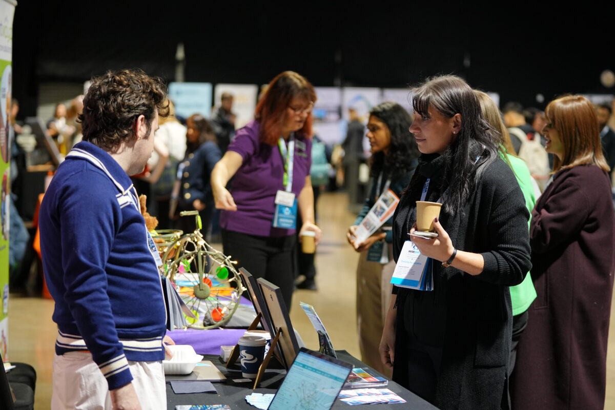 Several people stand at a table at a conference or exhibition, with a colorful, eclectic display in the foreground.