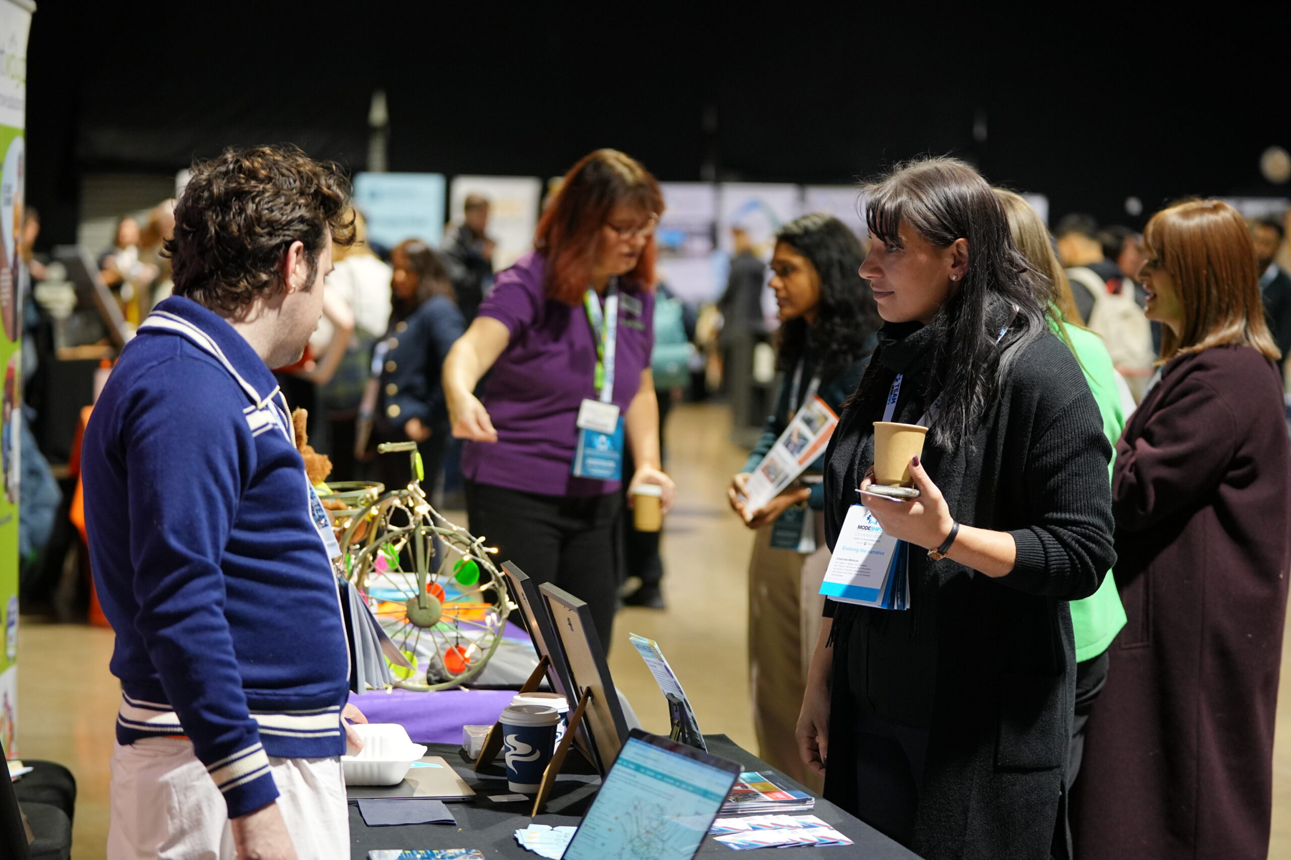 Several people stand at a table at a conference or exhibition, with a colorful, eclectic display in the foreground.