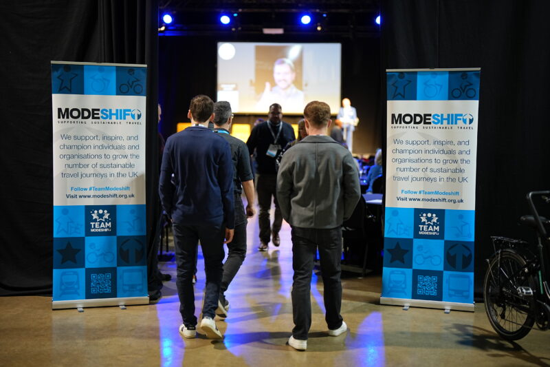Attendees walk towards a stage with a speaker projected on a screen, flanked by two ModeShift promotional banners that say "Supporting Sustainable Travel" and describe their mission.