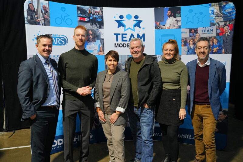 Six individuals pose in front of a blue backdrop with the "TEAM MODESHIFT" logo and various images related to cycling and transportation.